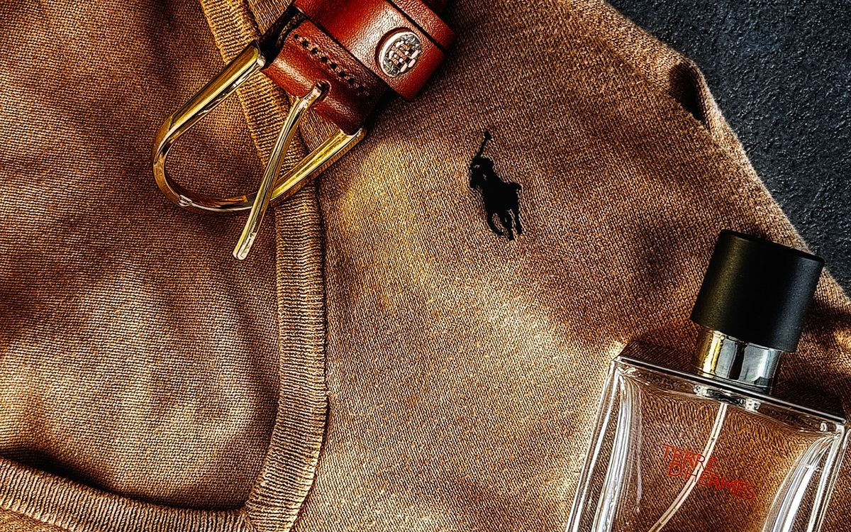 An evening flat-lay — a brown leather belt with a brass buckle laid across a camel knit, beside a small fragrance bottle