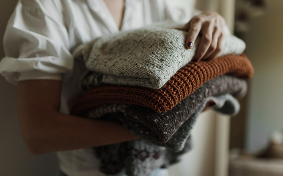 A stack of folded knitwear in cream, rust, and speckled grey, held in both arms